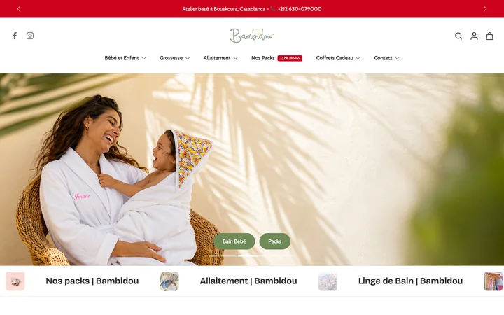 The Bambidou homepage on a desktop screen: a red announcement bar, the script wordmark above a French product navigation, and a wide photograph of a mother and child in white bathrobes in a wicker chair, above a row of category cards.